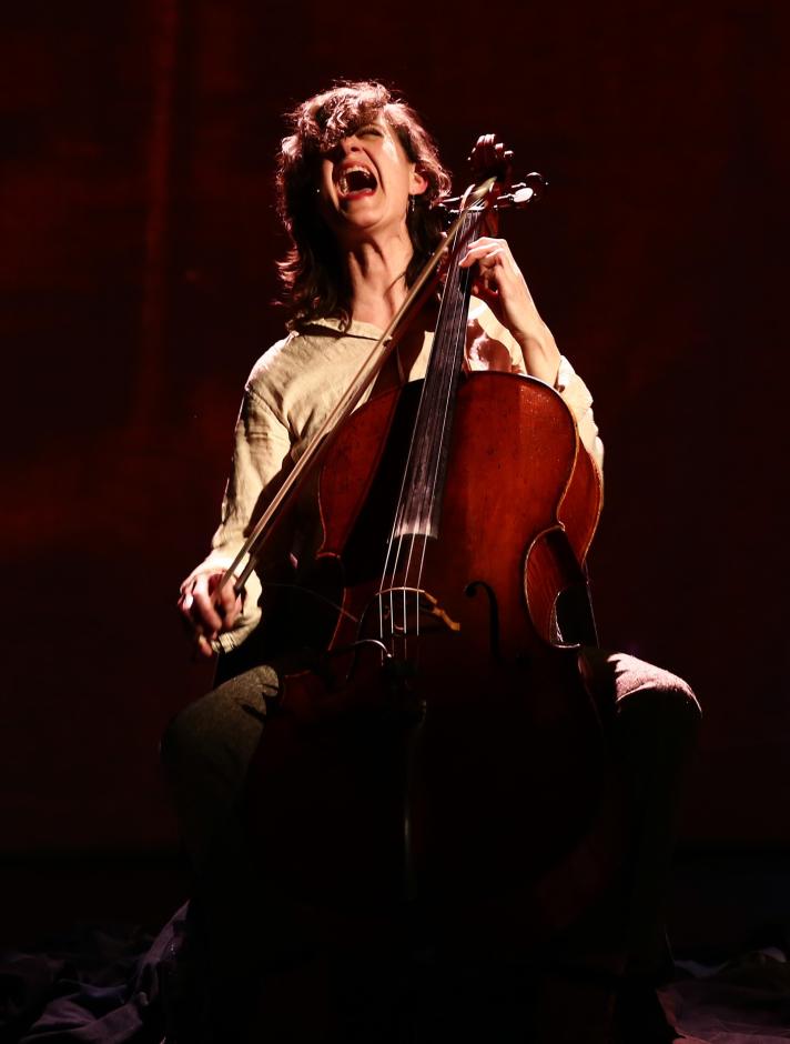 Woman playing cello and screaming wearing a beige blouse.