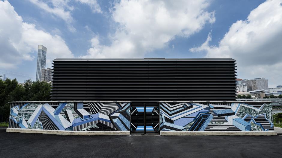 Street-level view of ventilation structure with blue, gray, white, and black mosaic artwork featuring a geometric design on the façade.
