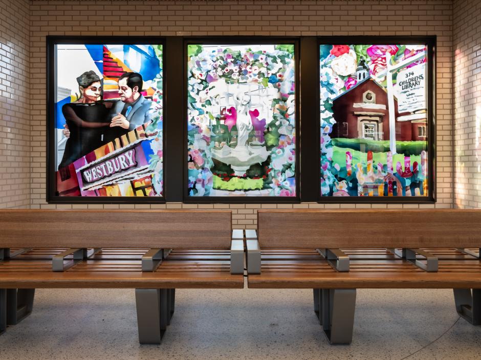 Interior view of a station waiting room with glass artwork in the background. Artwork panels depict people dancing, a garden, and a local library. 