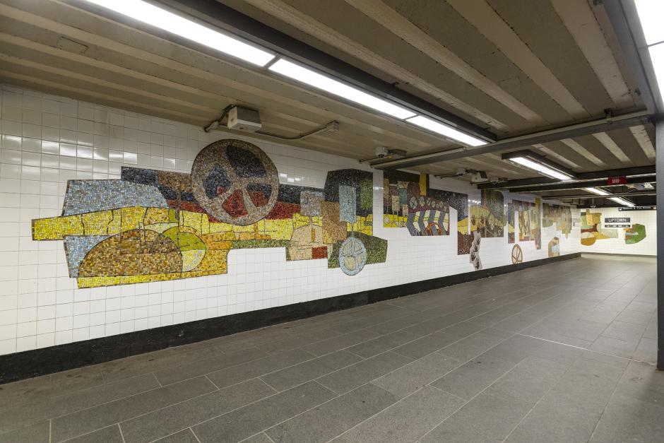 Station hallway with mosaic art on the wall. Mosaic artwork features abstract shapes and wheels.