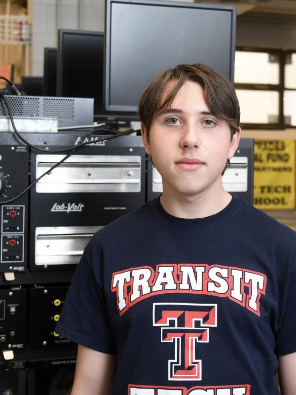 A young man wearing a Transit Tech shirt looks at the camera. Industrial equipment is visible behind him.