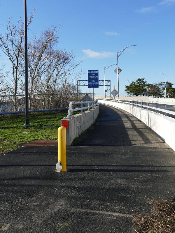 A bike lane next to the entrance to a bridge. The bike lane has grass on one side, and a white concrete barrier on the other. 