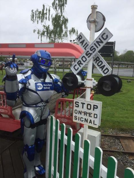 A person in a robot costume stands next to a road sign that says "Railroad Crossing"